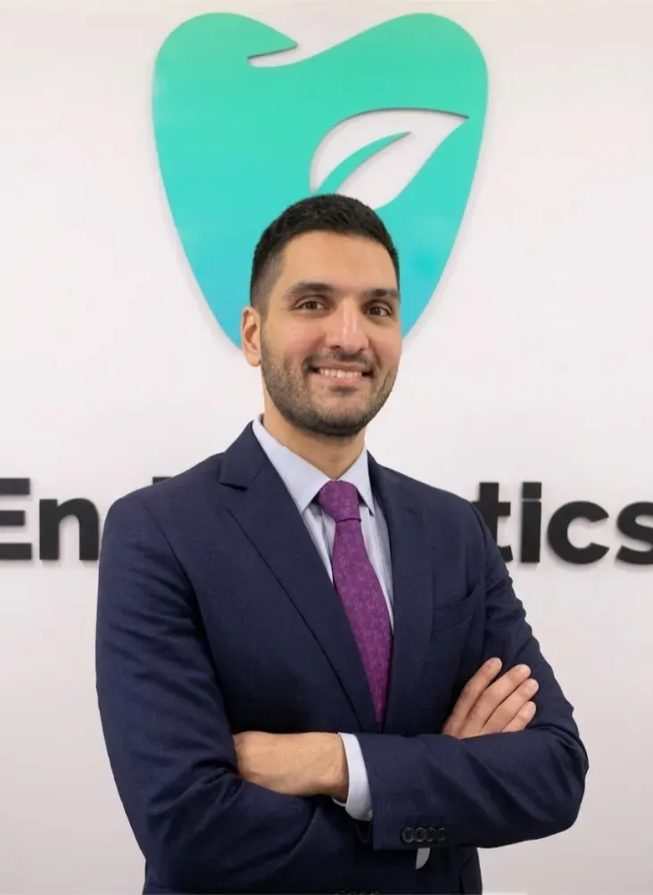 Professional headshot of a man in a navy suit and purple tie with arms crossed, standing in front of a teal tooth-shaped logo with partial text reading 'En...tics'.