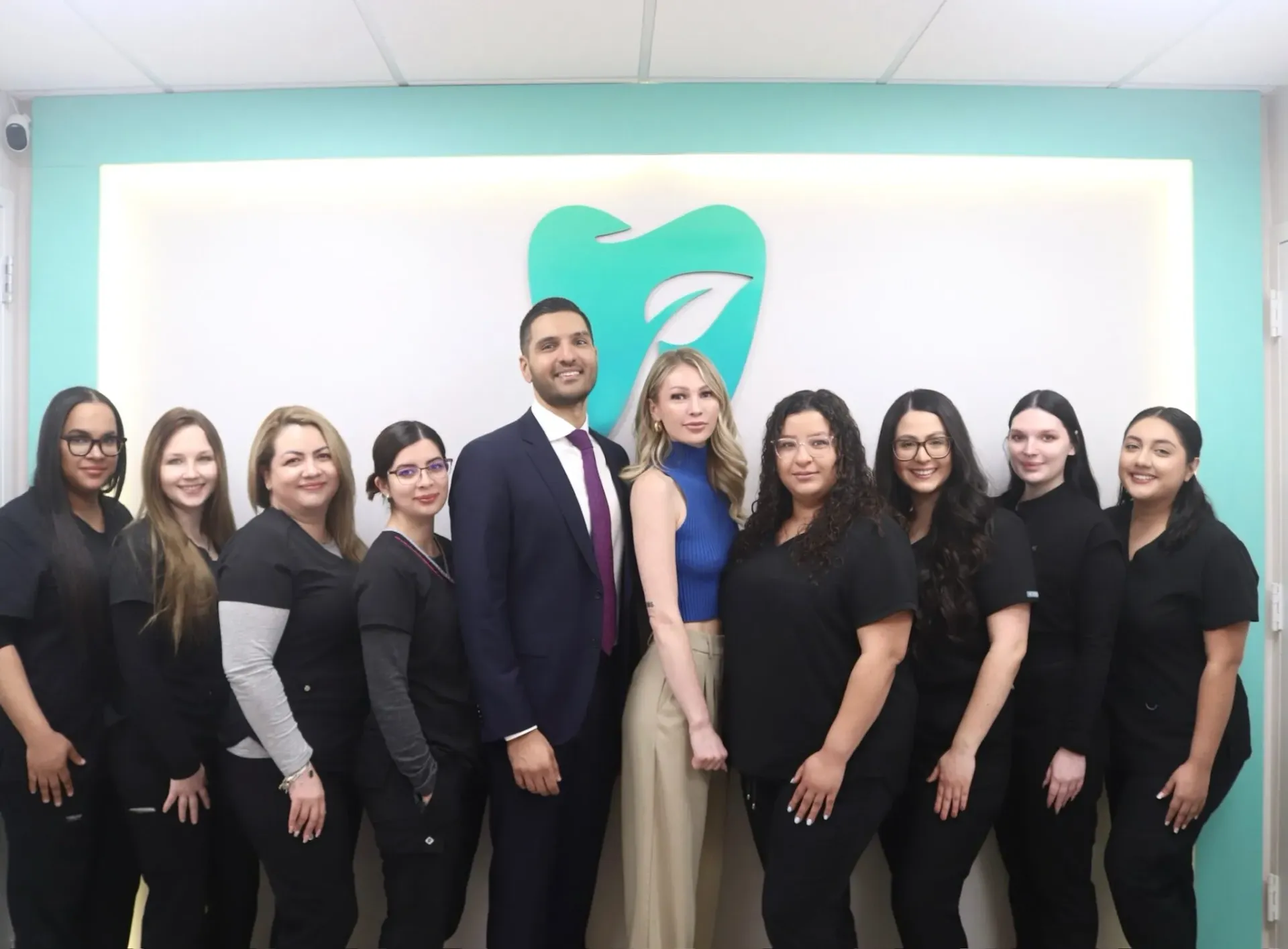 A group of nine dental office staff members posing in front of a teal wall featuring a prominent 3D tooth-with-leaf logo; staff wear black scrubs while two central figures wear business attire.