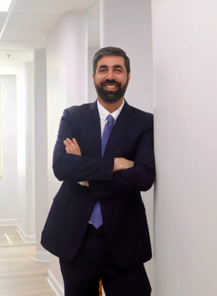 Professional headshot of a smiling man with a beard wearing a dark navy suit and purple tie, arms crossed, leaning against a white wall in a bright office hallway.