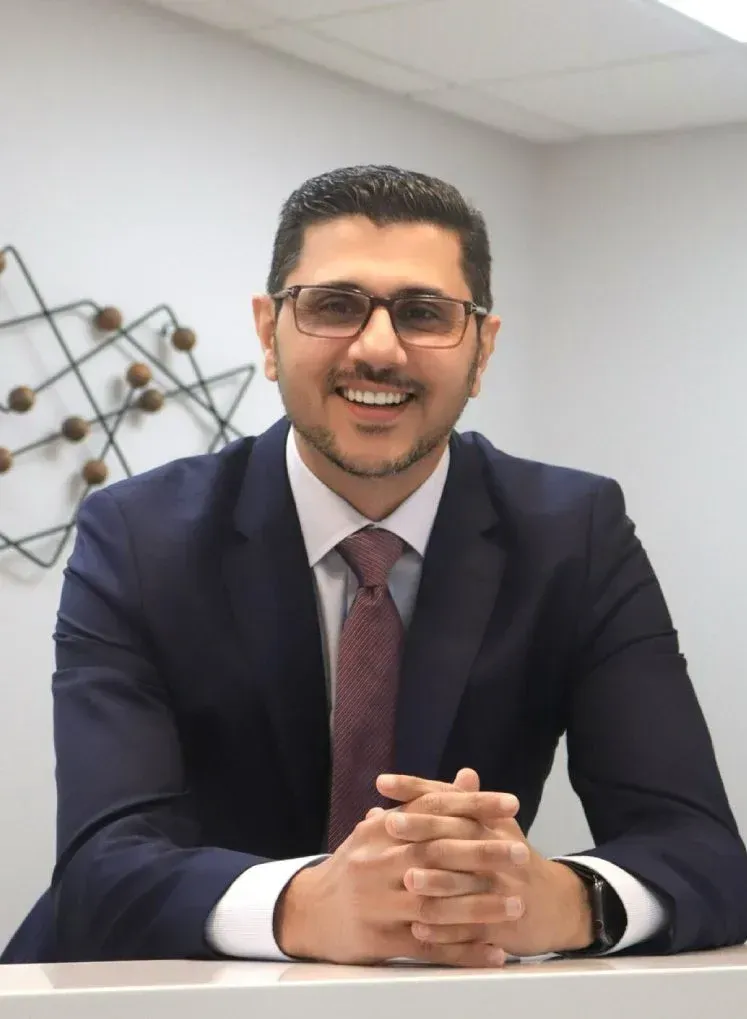 Professional headshot of a smiling man wearing glasses, a navy suit, and a dark red tie, seated at a desk with hands clasped