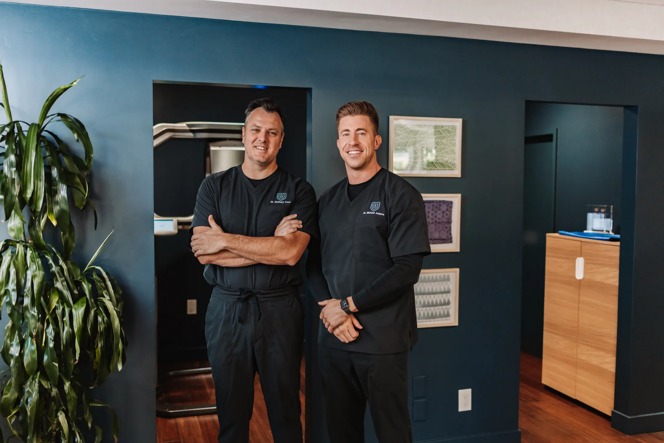 Two male dental professionals in black scrubs labeled 'Dr. Zachary Freer' and 'Dr. Michael Adams' smiling in the office hallway against a teal accent wall. Professional, well-lit portrait suitable for team or about sections.