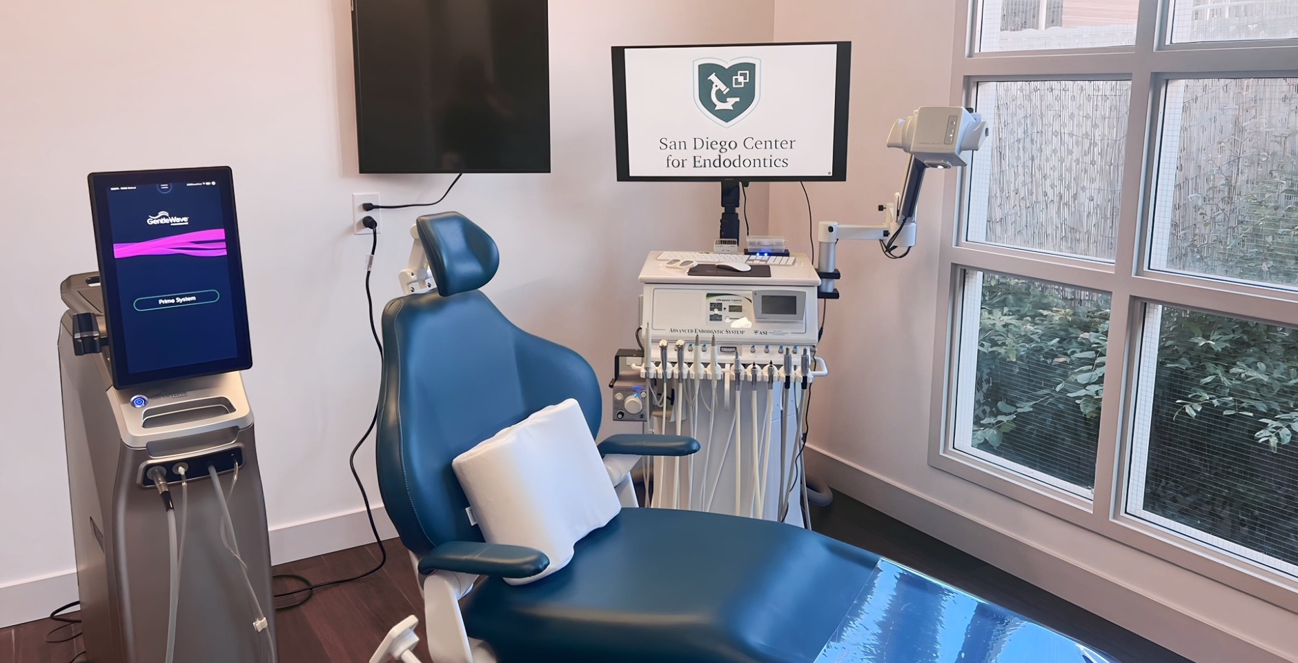 Modern endodontic treatment room featuring a teal dental chair with pillow, GentleWave Prime System device on the left, Advanced Endodontic System cart, microscope arm, and a monitor displaying the 'San Diego Center for Endodontics' logo.