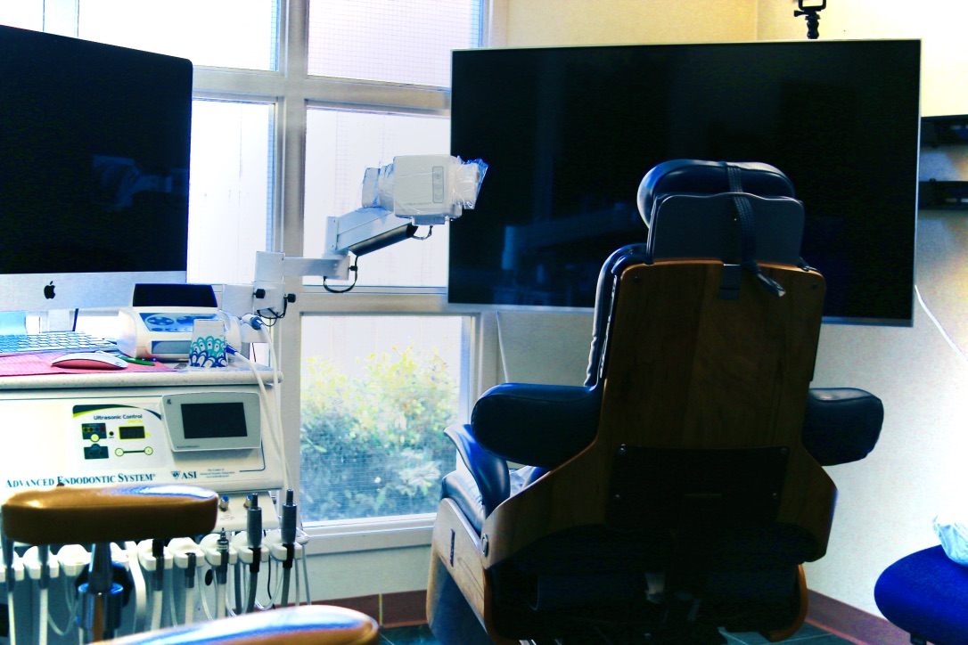 Endodontic operatory showing a navy dental chair with wooden back, 'Advanced Endodontic System – ASI' cart, wall-mounted microscope, and two monitors, with grid windows in the background.