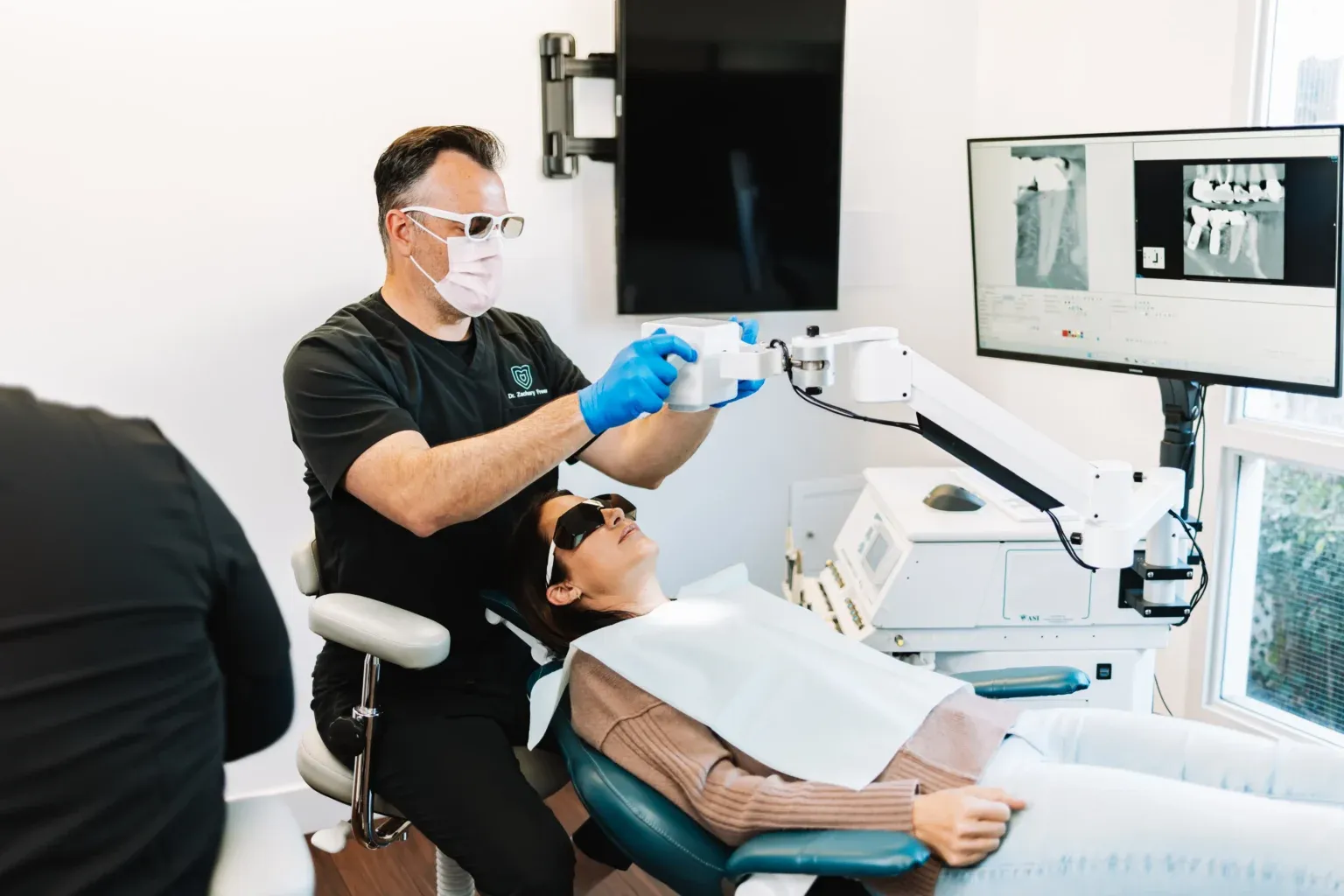 Interior view of a modern dental treatment area with dental chairs, equipment arms, teal accent walls, wood-look flooring, and a large potted plant near an X-ray machine alcove.