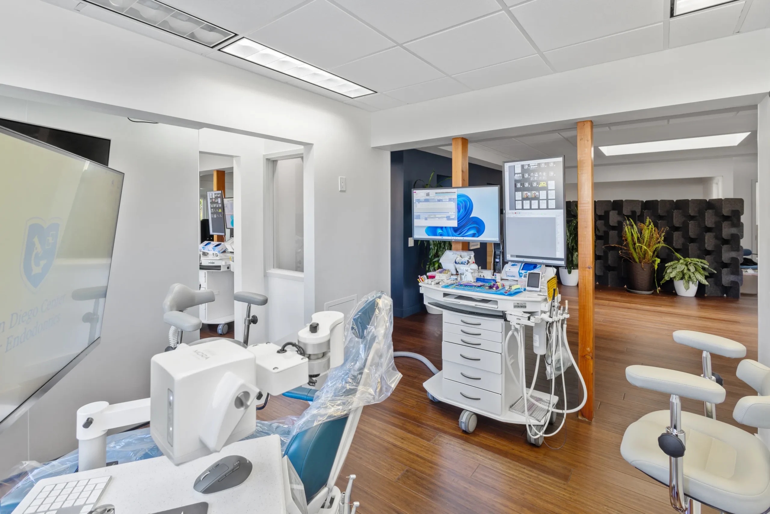 Modern dental treatment room with a dental chair, overhead equipment, dual monitors displaying Windows 11, and a partially visible logo reading 'San Diego Center for Endodontics' on the wall; professionally photographed. Well-equipped and contemporary clinical space.