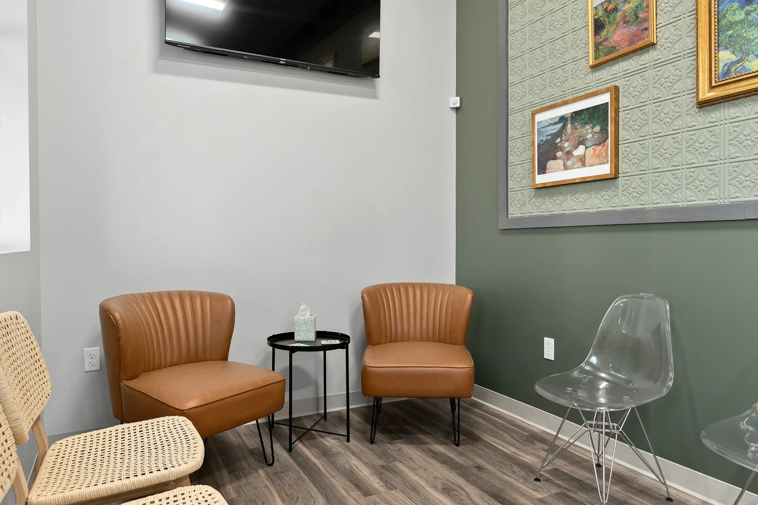 Modern dental or medical office waiting room featuring tan leather chairs, a clear acrylic chair, a small black side table, and framed artwork on a sage green textured accent wall with a wall-mounted TV. High-resolution, well-lit interior photo showcasing a welcoming patient space.