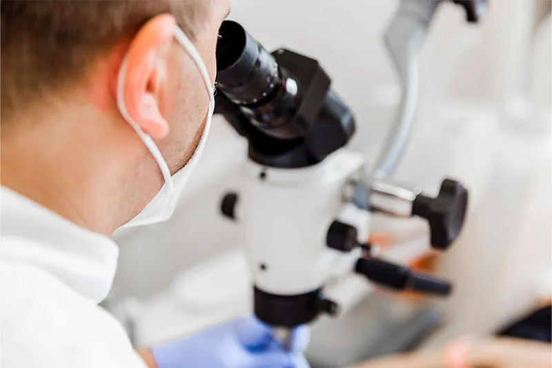 A masked dental professional in a white coat looks through a dental operating microscope, shown in a close-up side profile with shallow depth of field.