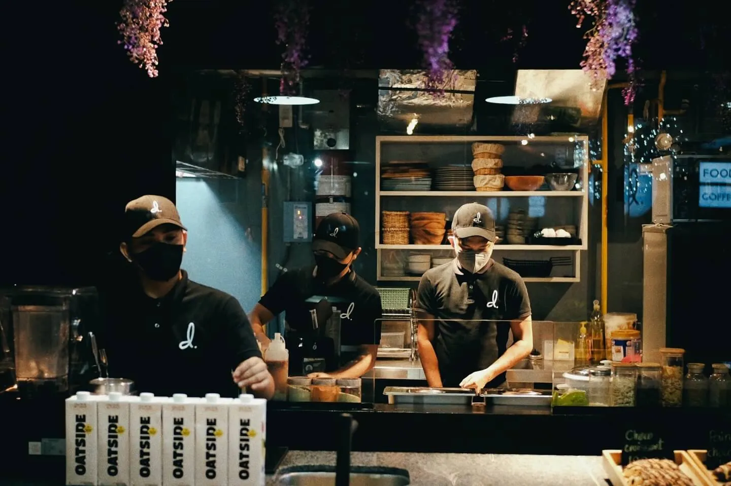 Three café staff members in matching black uniforms with a stylized 'd.' logo and caps working behind an open kitchen counter