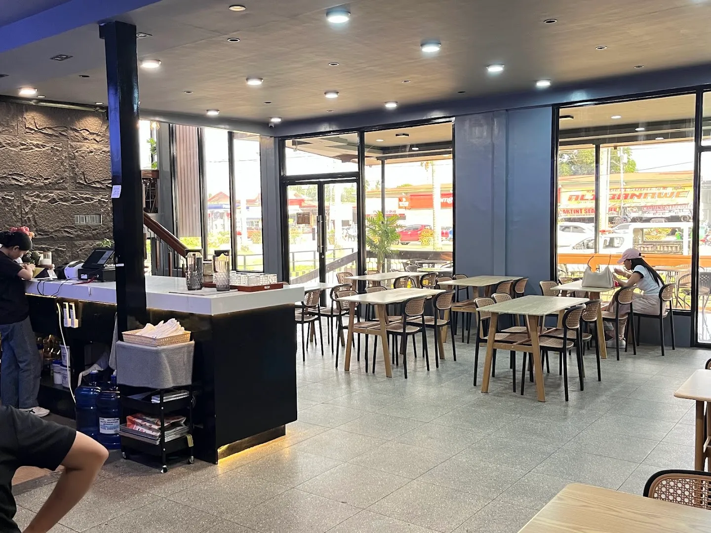 Bright, spacious interior of Daleachious Cafe showing the counter/POS area on the left and multiple dining tables with rattan-style chairs, large floor-to-ceiling windows revealing a street view outside.