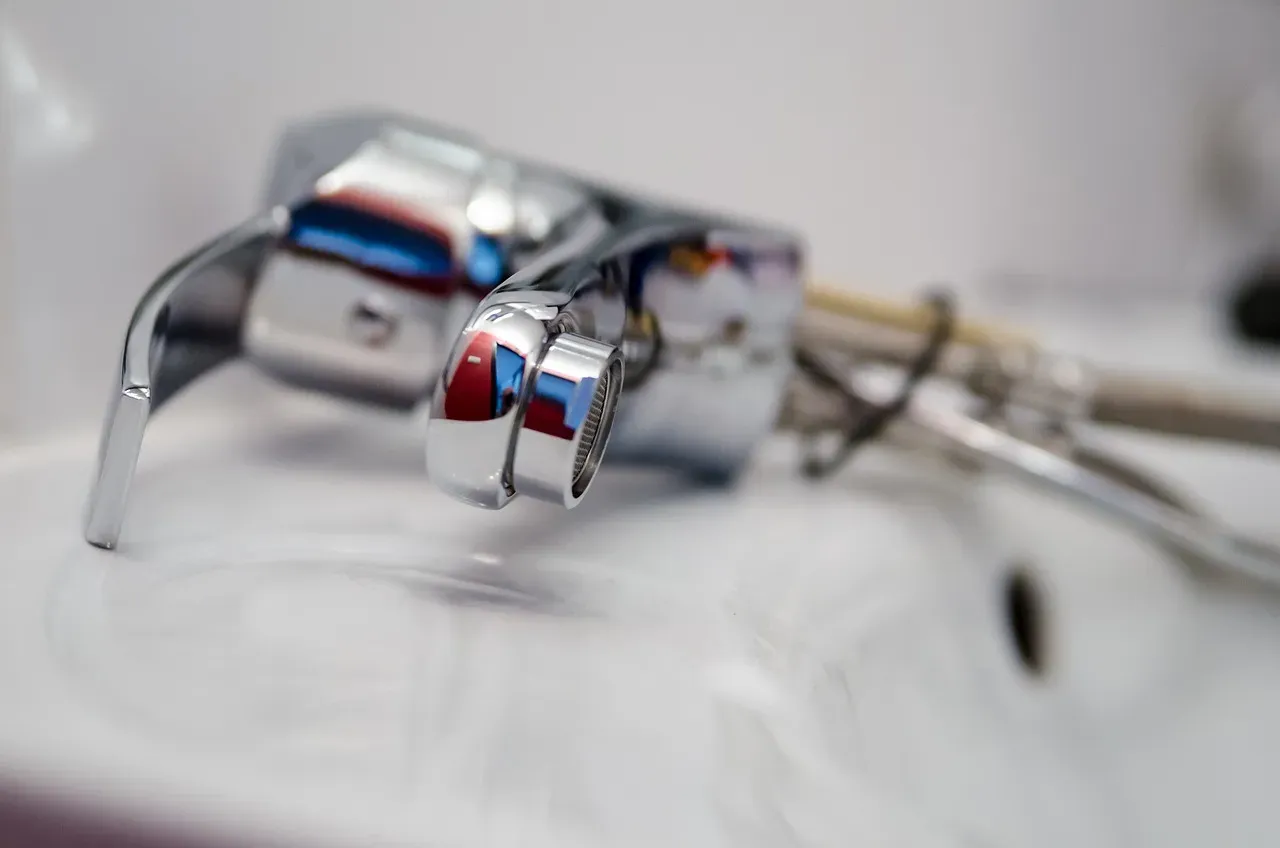 A shallow depth-of-field close-up of a chrome bathroom faucet being installed or repaired, with plumbing tools visible in the blurred background.
