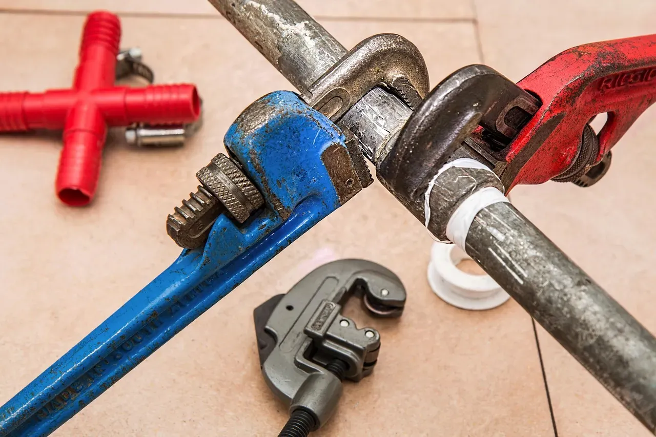 A close-up of plumbing tools including a blue pipe wrench and a red Ridgid pipe wrench gripping crossed metal pipes on a tile floor.