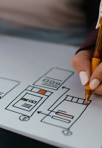 A close-up of a hand holding a gold pen sketching UI/UX wireframe layouts on white paper, with orange accent elements visible in the wireframe boxes.