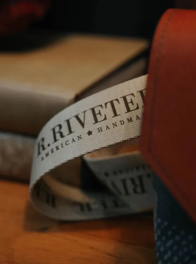Close-up product/branding shot of a fabric ribbon label reading 'R. RIVETER. AMERICAN ★ HANDMA[DE]' wrapped around leather goods