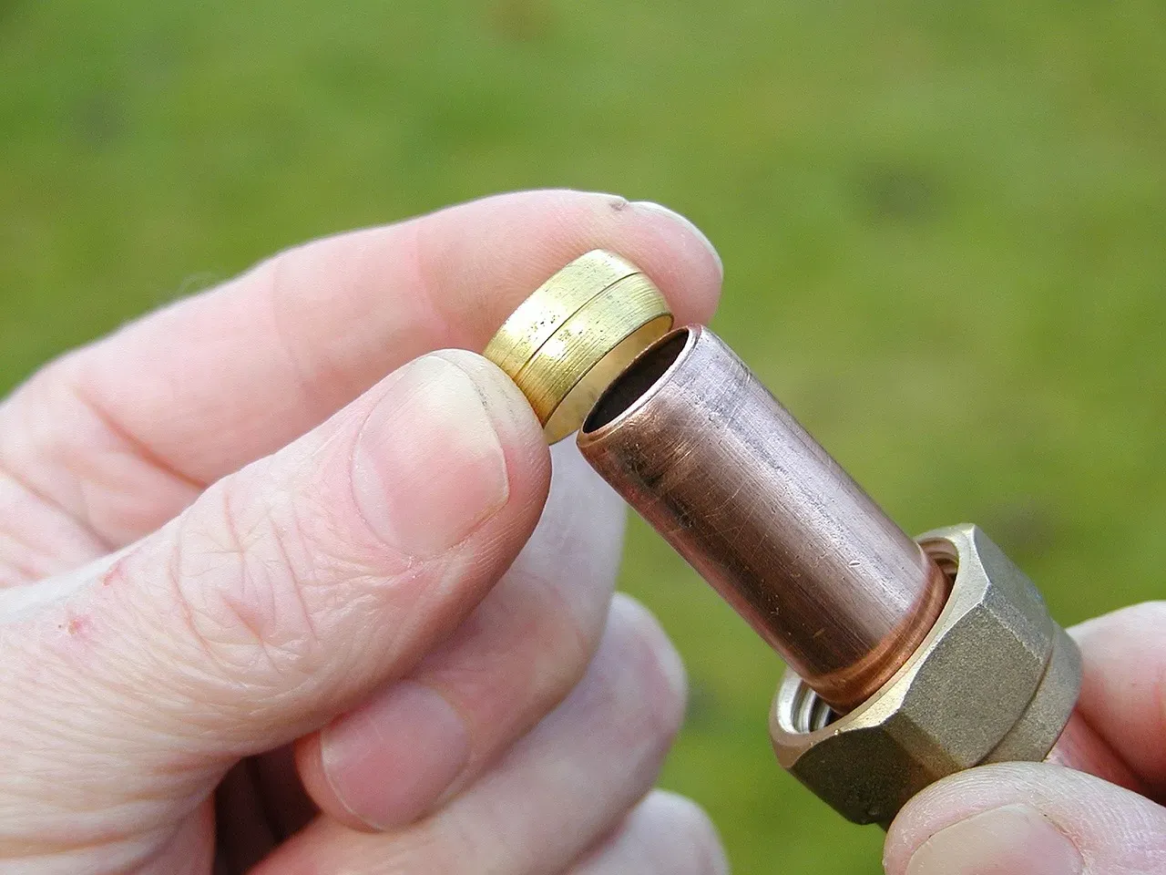 Close-up of two hands holding a copper pipe being fitted into a brass compression fitting, photographed against a blurred green grass background.