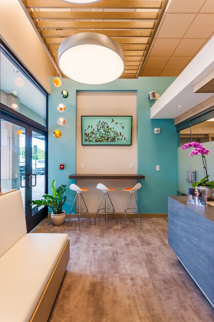 Professional interior photo of the Caswell Orthodontics waiting/reception area featuring teal accent walls, a large circular pendant light, colorful globe pendant lights, three white-and-orange bar stools at a counter, a framed bird artwork, orchid flowers, and a plant.