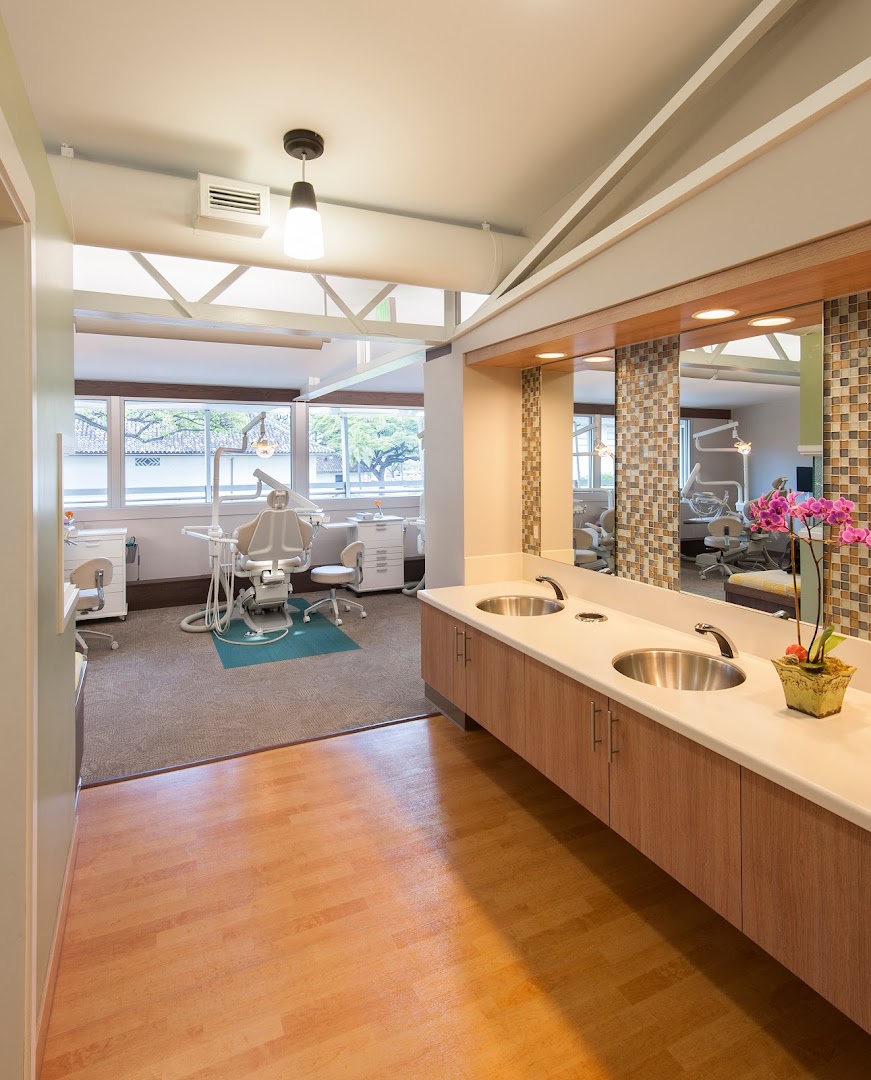 Professional dental office interior showing a treatment bay with a dental chair and equipment in the background, and a double-sink vanity station with mosaic tile backsplash and orchid arrangement in the foreground.
