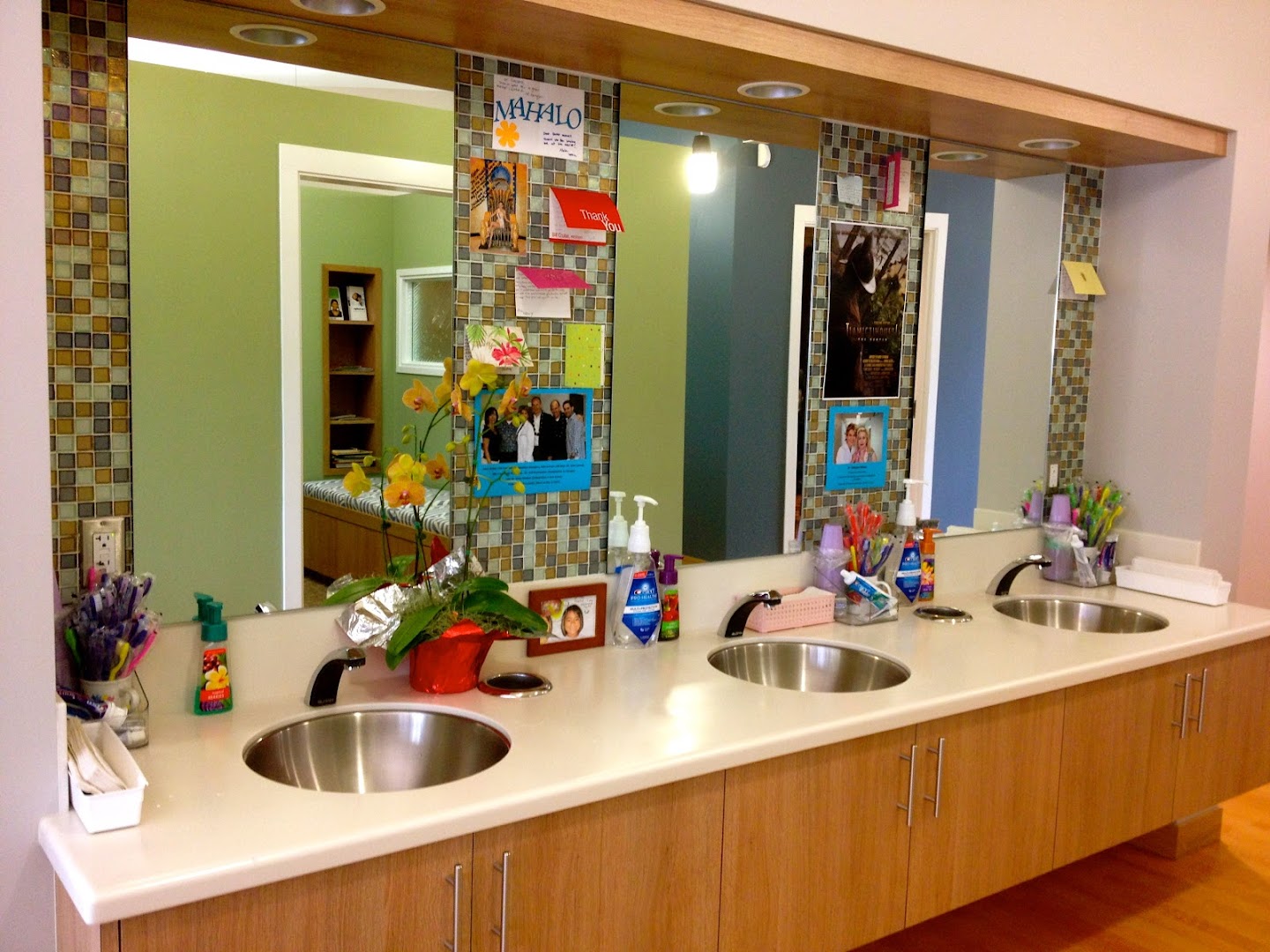 A dental office sink/vanity area with three round stainless steel sinks, mosaic tile-framed mirrors, orchid flowers, toothbrushes, and patient notes including visible text 'MAHALO' and 'Thank You' cards.