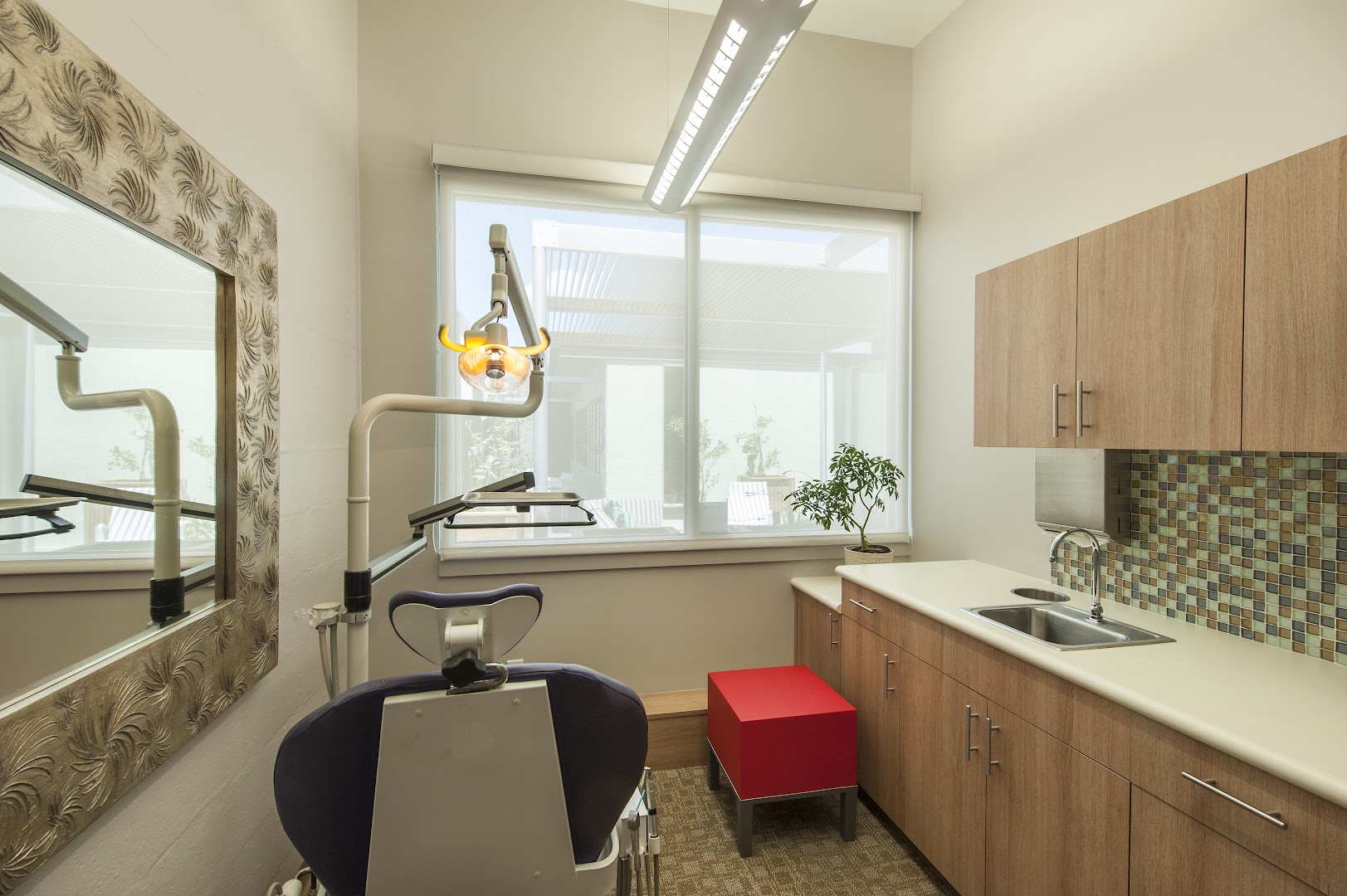 Professionally photographed private dental treatment room featuring a blue dental chair, overhead exam light, decorative ornate mirror, mosaic tile backsplash, wood cabinetry, and a small bonsai plant on the counter.
