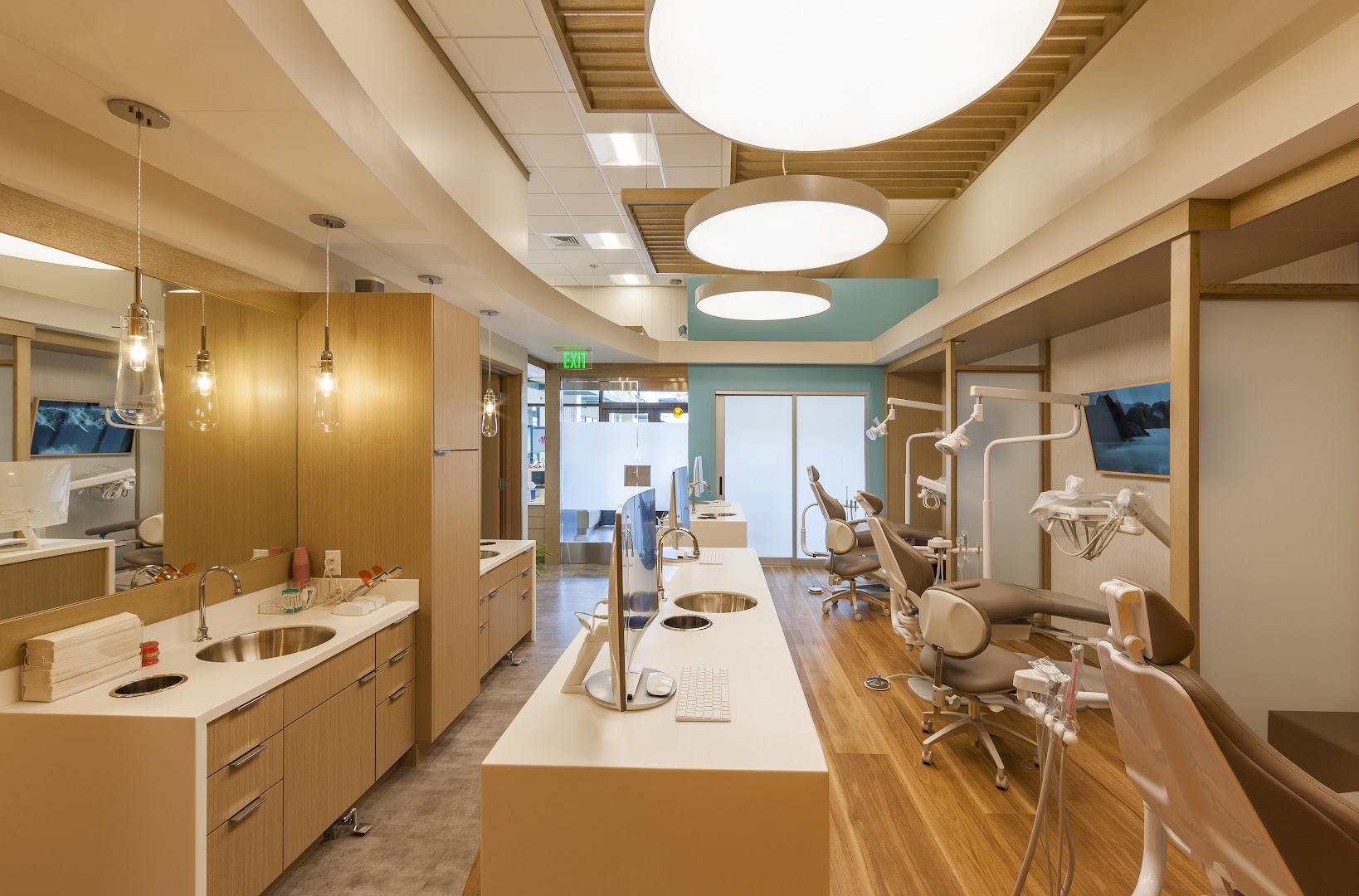 Professional interior photograph of Caswell Orthodontics open-bay treatment area showing multiple dental chairs, iMac workstations, warm wood cabinetry, pendant lighting, and large circular ceiling lights with a teal accent wall visible in the background.