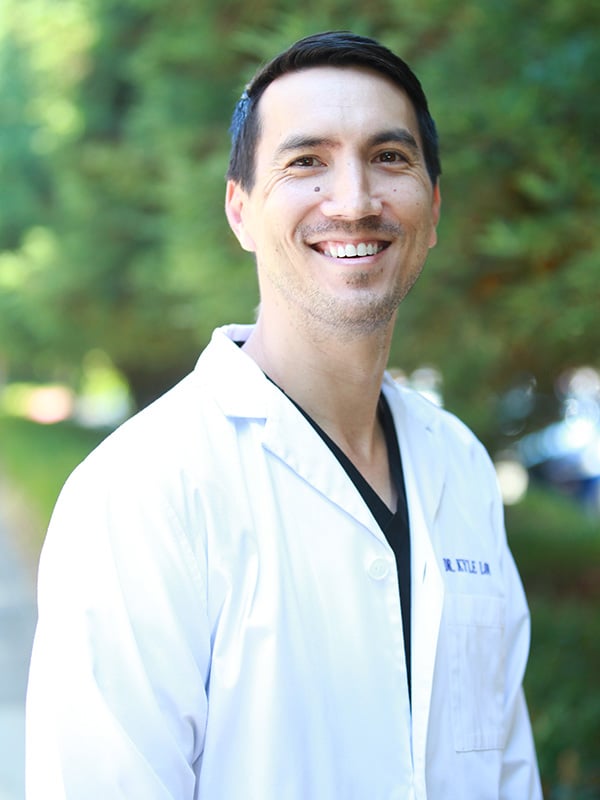 Duplicate outdoor portrait of the same smiling male doctor in a white lab coat with 'Dr. Kyle L...' embroidered on the chest, identical composition and quality to the previous image. Blurred green foliage background.