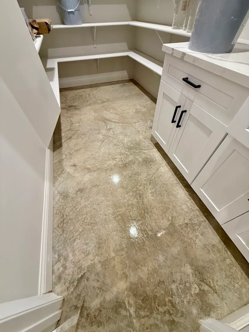 A narrow utility or laundry room with a polished concrete or epoxy floor in a warm beige/taupe tone with a subtle marbled texture, flanked by white cabinetry and shelving.