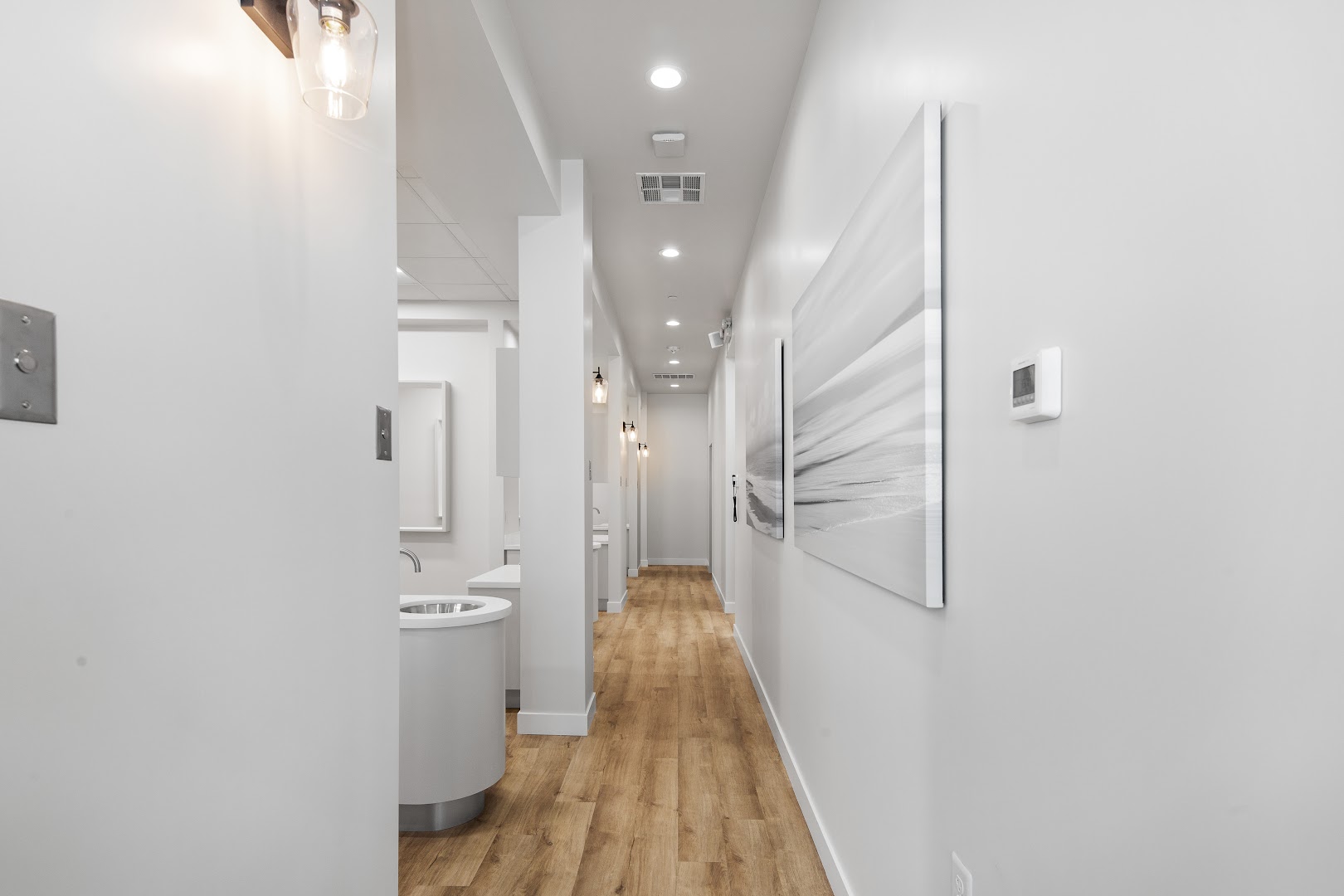 Clean, white dental office hallway with warm wood flooring, pendant lighting, abstract black-and-white wall art, and treatment room entrances visible along the corridor. High-quality architectural interior shot conveying a modern, upscale clinic environment.