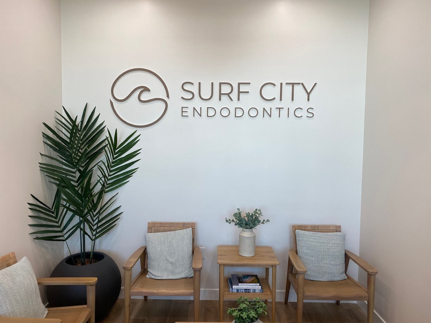 Wall-mounted 3D logo reading 'SURF CITY ENDODONTICS' with a wave-circle icon, displayed above a modern waiting area with wooden chairs, a palm plant, and neutral decor. High-quality, well-lit interior branding shot.