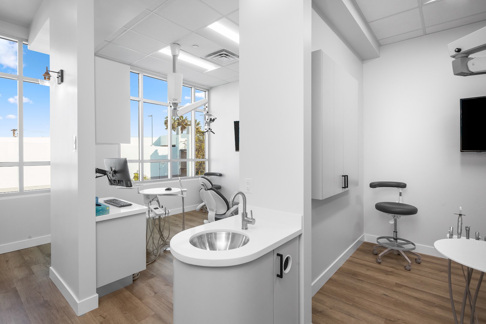 Modern dental treatment room with a dental chair, overhead light, computer workstation, stainless steel sink, and large windows overlooking palm trees and blue sky. Sharp, professionally photographed interior showcasing state-of-the-art equipment.