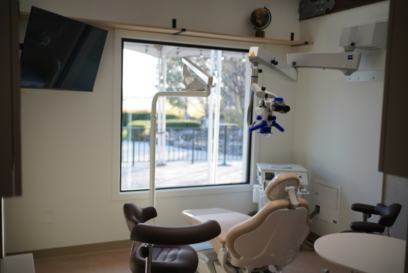 Modern endodontic treatment room featuring a dental chair, ASI dental unit, operating microscope, and stylish pendant lighting with dark wood beam accents. High-quality interior photo showcasing a well-equipped, aesthetically designed operatory.