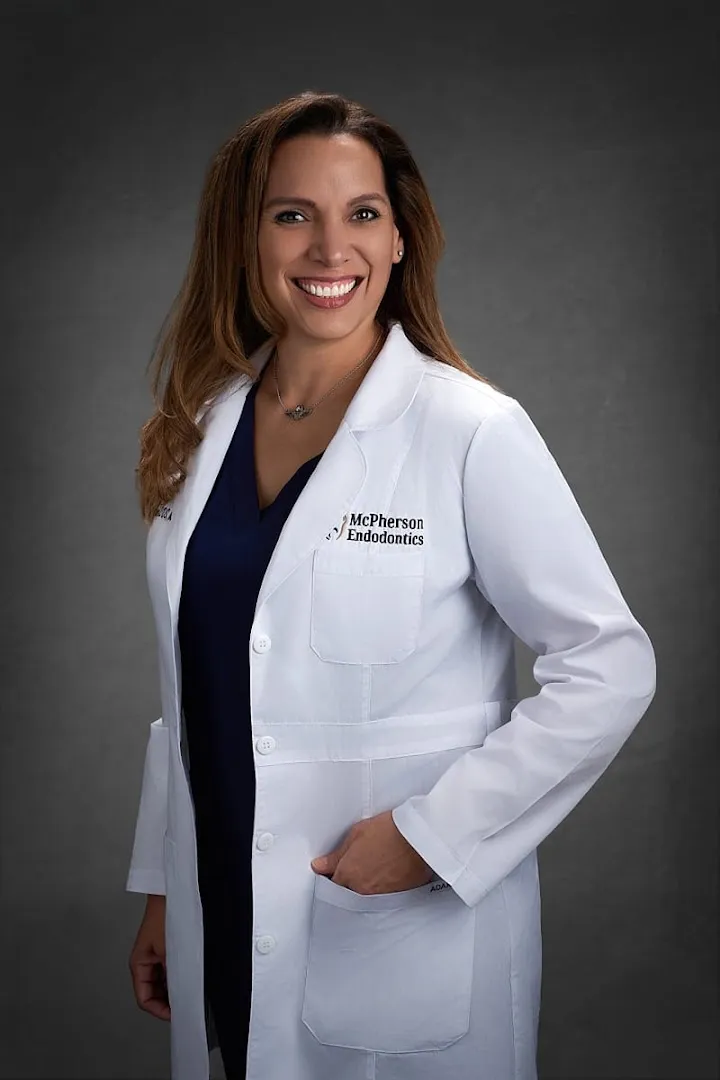 Professional full-body headshot of a smiling female doctor in a white lab coat clearly embroidered with 'McPherson Endodontics,' wearing a navy scrub top