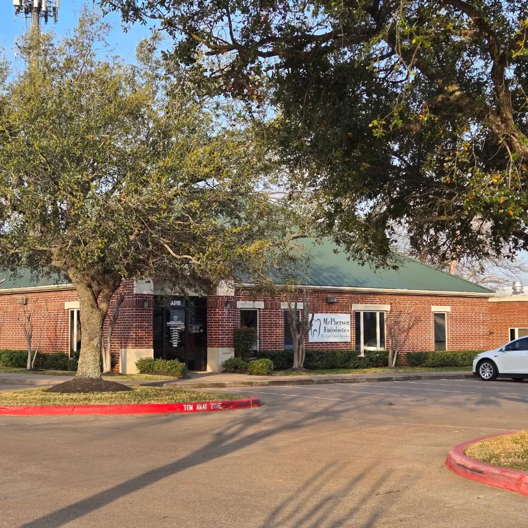 Exterior daytime photo of the McPherson Endodontics brick office building at 3318, with signage on the facade and a large oak tree.