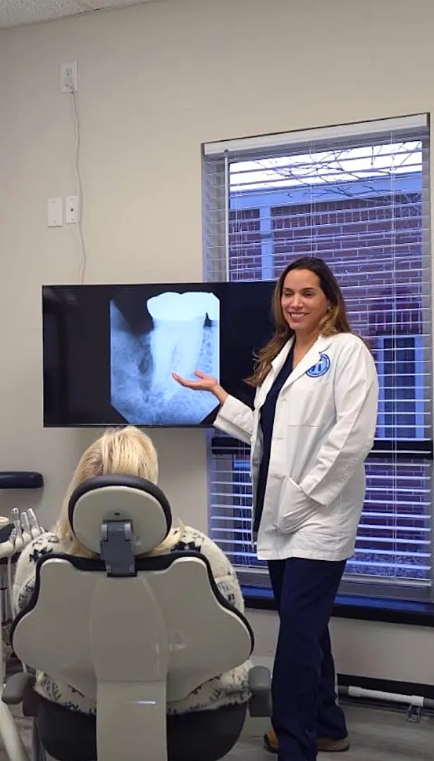 A female dentist in a white lab coat stands beside a dental chair, gesturing toward an X-ray displayed on a wall-mounted monitor in a clinical treatment room.