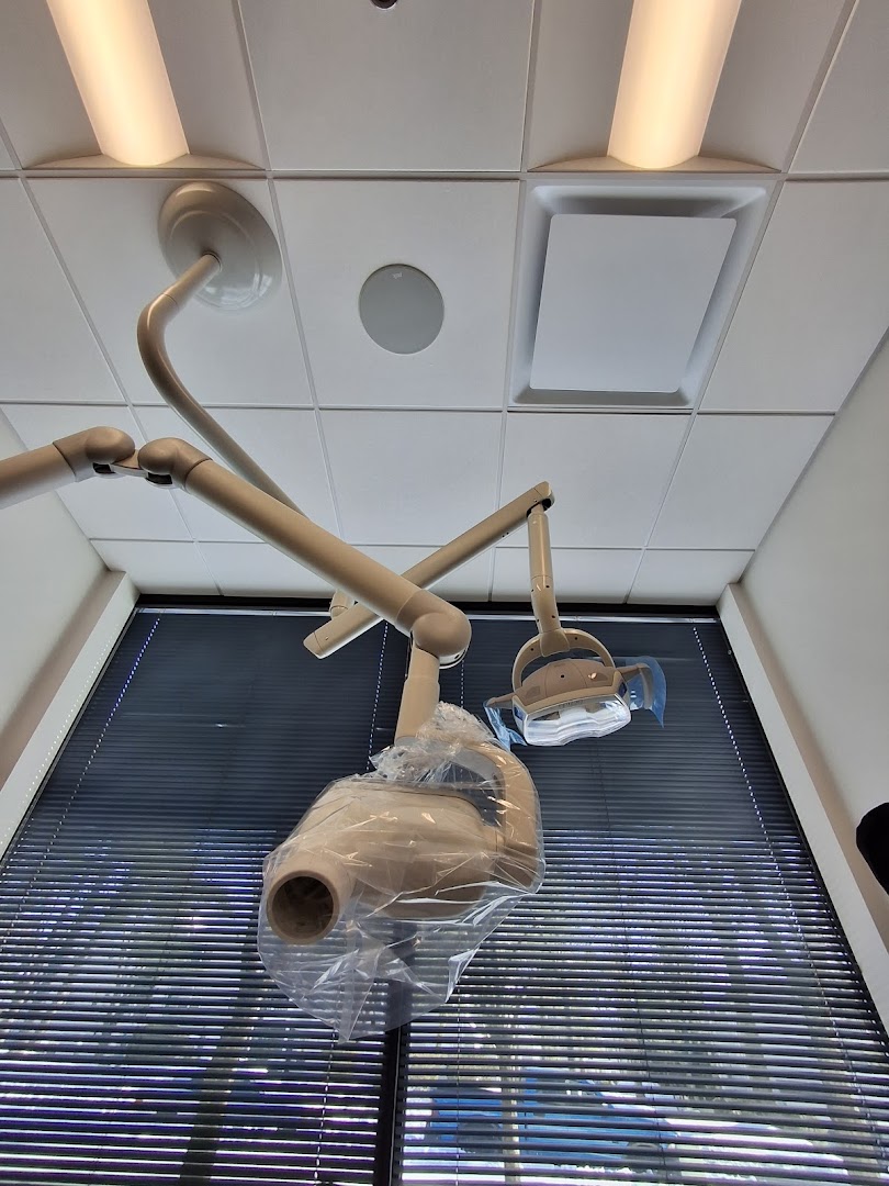 Upward-angle shot of dental operatory ceiling equipment including an articulating arm, overhead light, and suction apparatus wrapped in plastic barrier, with window blinds visible in the background.