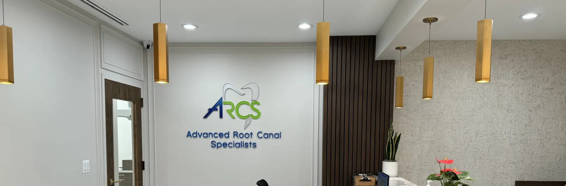 Panoramic interior shot of the ARCS Advanced Root Canal Specialists reception area showing the branded wall logo, gold cylindrical pendant lights, dark wood slat accent wall, and a partial view of the marble reception desk with a plant and flowers. The image is wide and well-lit with a clean, modern aesthetic.