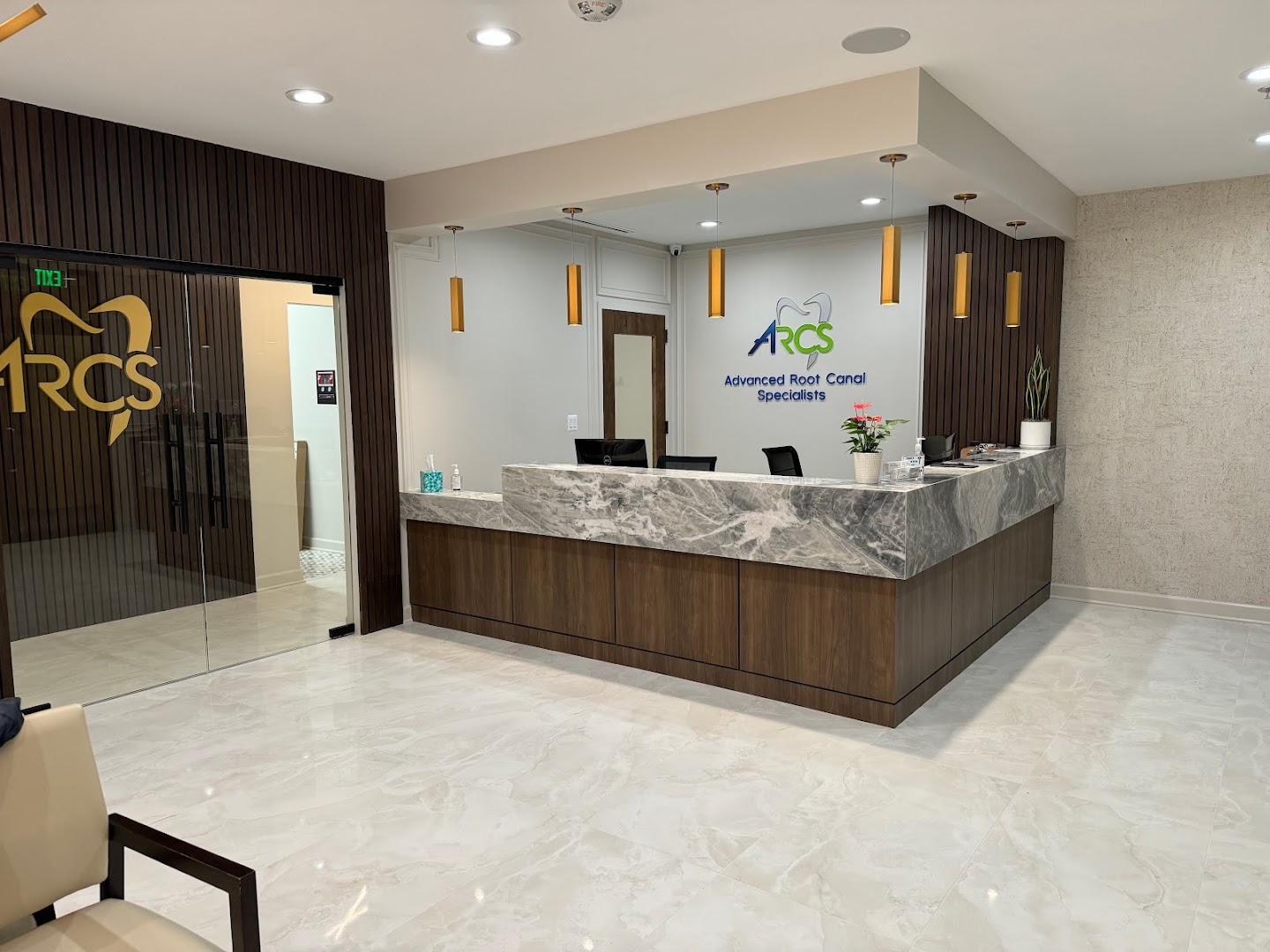 Interior shot of the ARCS Advanced Root Canal Specialists reception area showing the marble-topped front desk, ARCS logo on the back wall, gold pendant lights, and the branded glass entrance door.