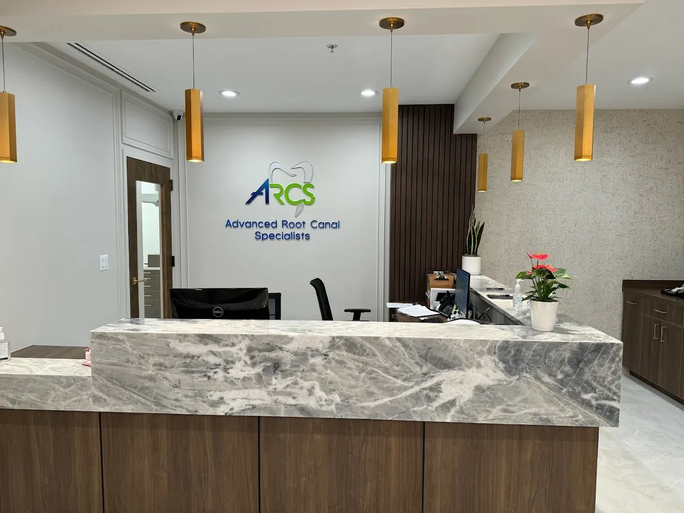 Panoramic interior shot of the ARCS Advanced Root Canal Specialists reception area showing the branded wall logo, gold cylindrical pendant lights, dark wood slat accent wall, and a partial view of the marble reception desk with a plant and flowers. The image is wide and well-lit with a clean, modern aesthetic.