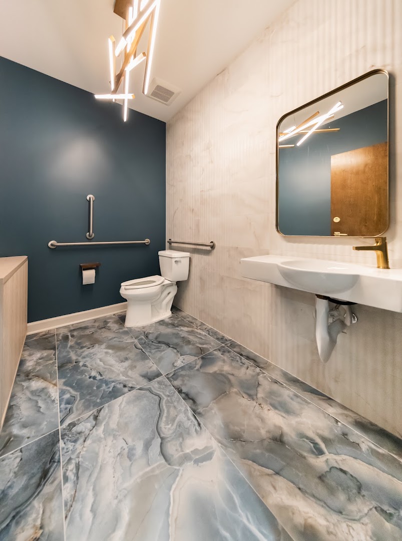 Stylish accessible restroom featuring dramatic blue-veined marble tile flooring, a navy accent wall, gold fixtures, a floating white sink, and a modern geometric chandelier.