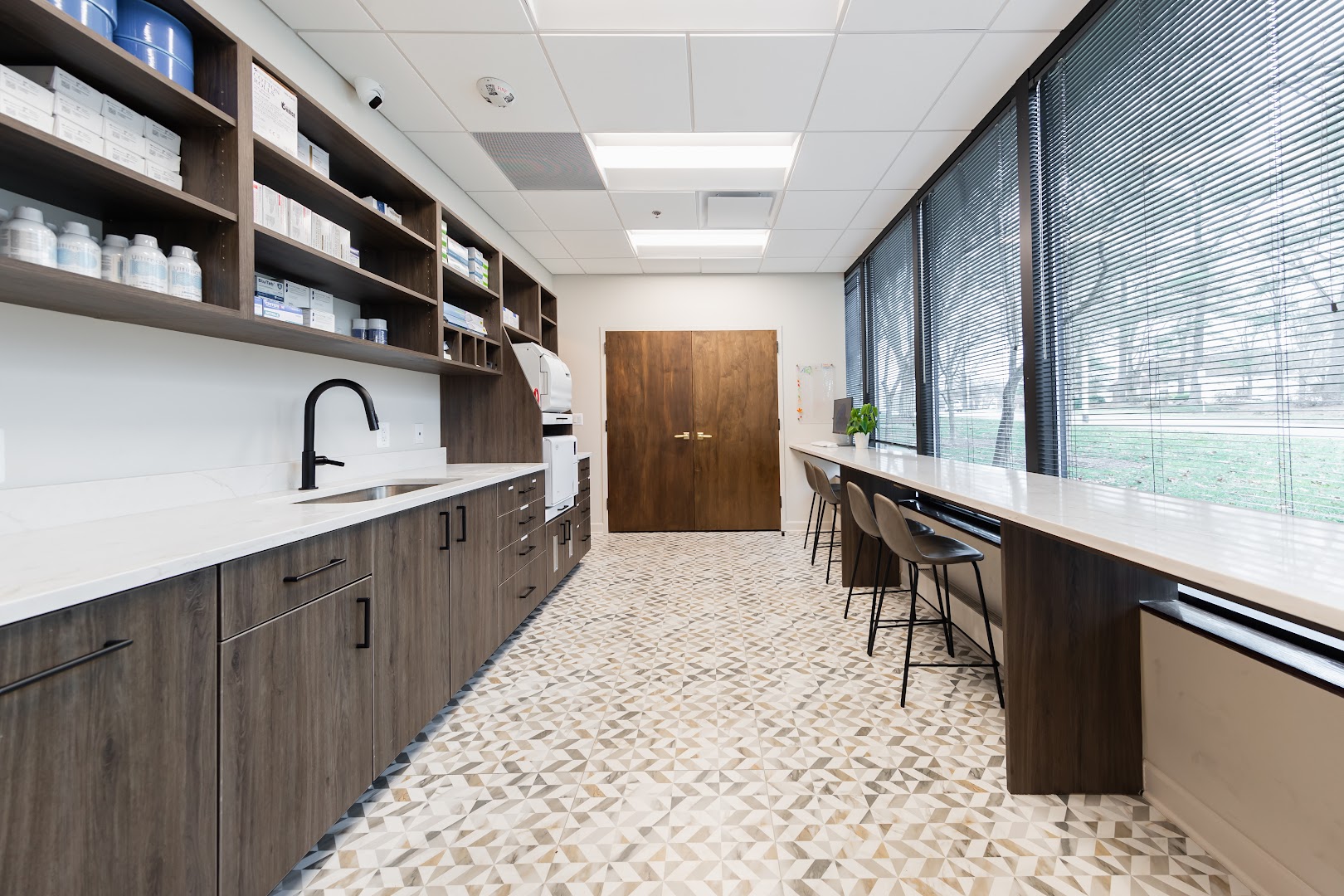 Professional shot of a well-organized dental sterilization or staff workroom with dark wood cabinetry, white quartz countertops, open shelving stocked with medical supplies, a black matte faucet, and a bar-height counter along large windows.