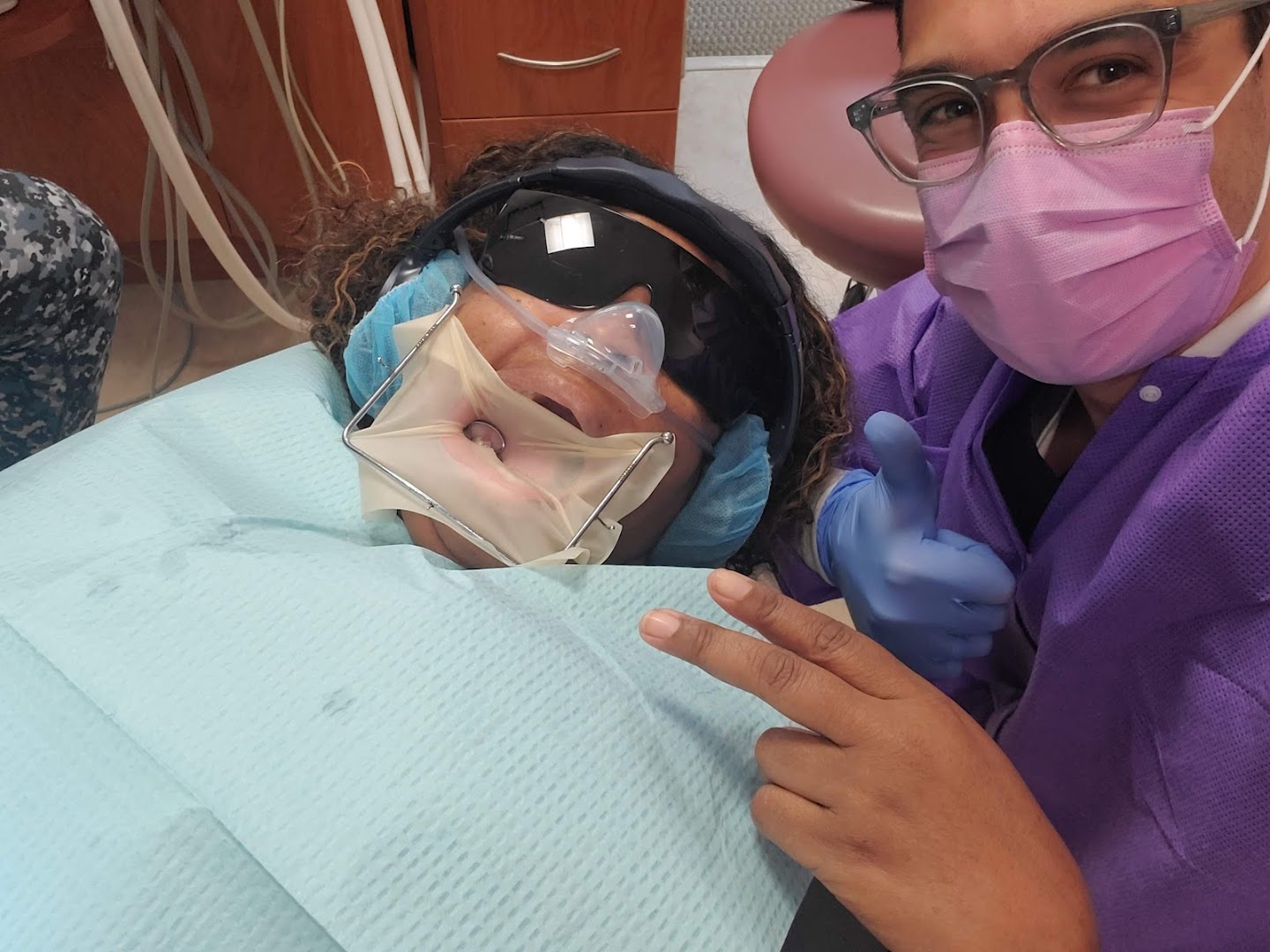 Selfie-style photo of a dental professional in purple scrubs and pink mask posing next to a patient in a dental chair who is wearing protective goggles and a rubber dam.